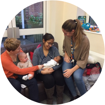 Two mothers sitting with their babies with a volunteer looking down at the youngest baby